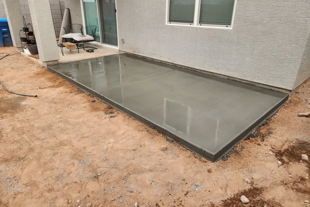 Freshly poured concrete patio slab along the back of a stucco house, with the yard still bare dirt